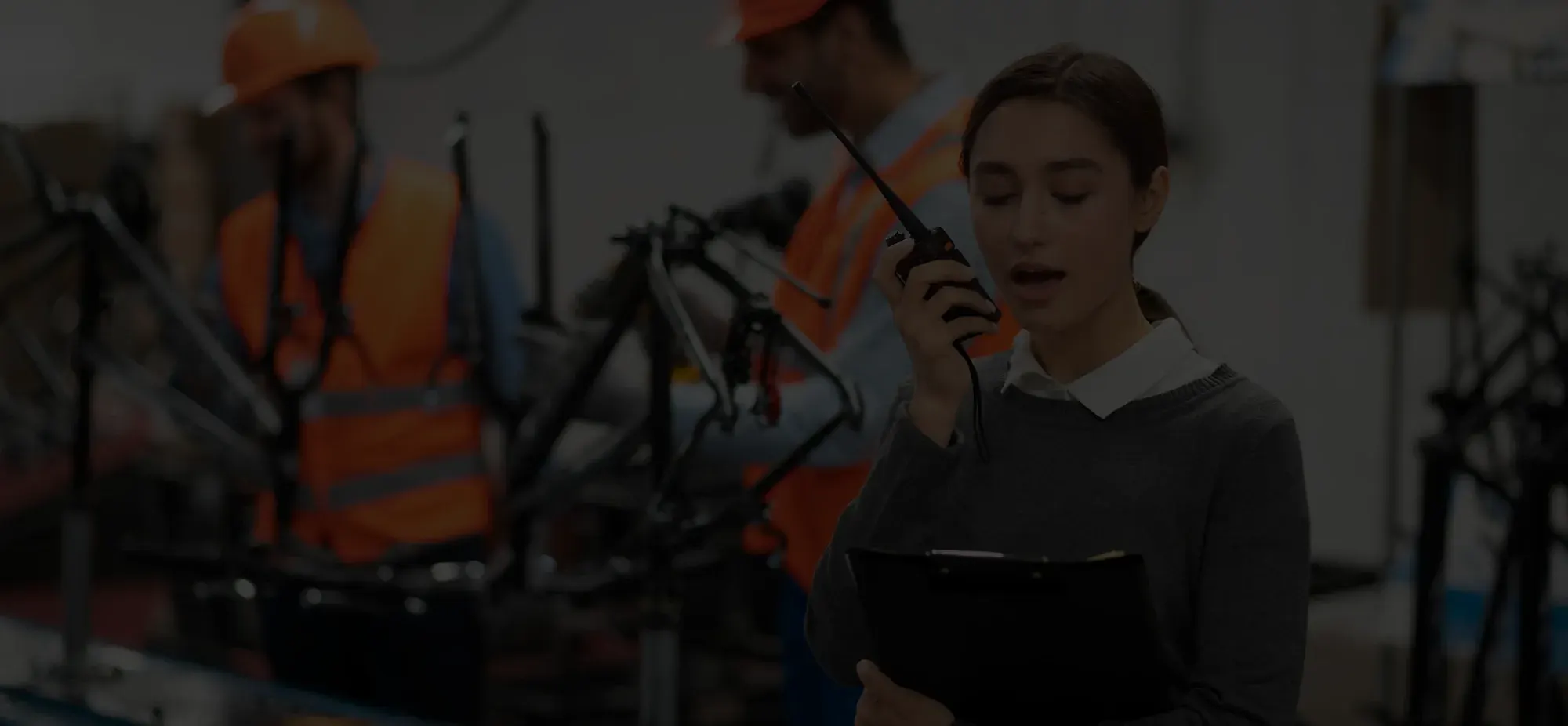 Woman holding a clipboard and speaking into a walkie-talkie in a factory setting with workers assembling bicycles in the background.