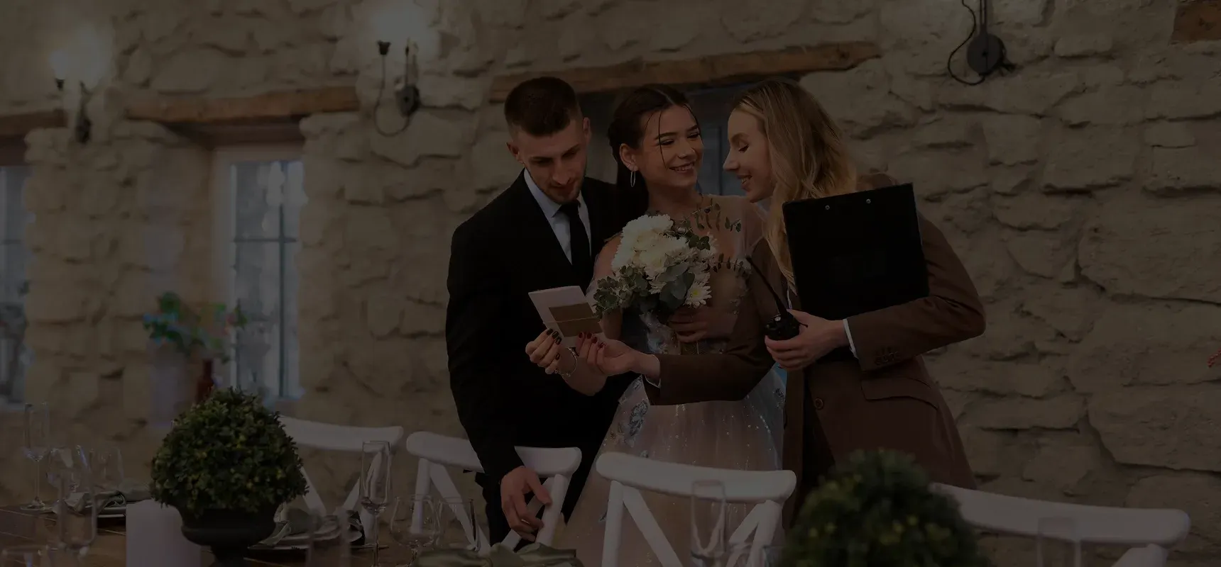 Bride holding a bouquet and smiling with groom and wedding planner reviewing a card in a rustic stone-walled room.