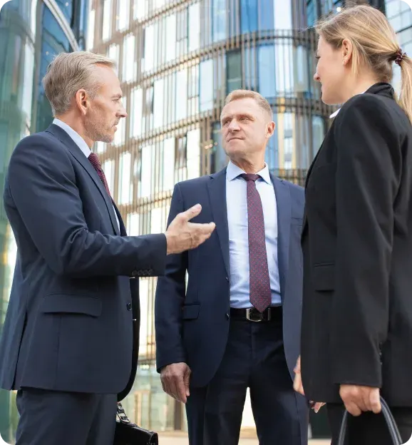 Three business professionals in suits having a discussion outside a modern glass office building.