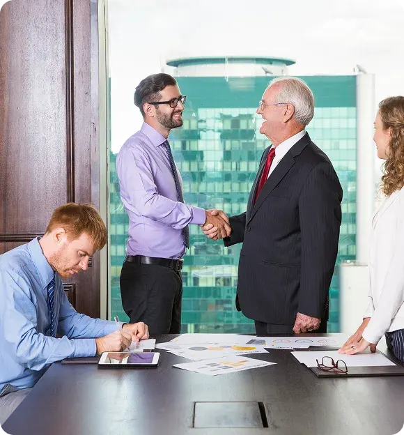 Two men in business attire shaking hands across a conference table with documents, a tablet, and two other colleagues present.