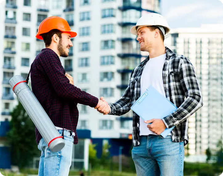 Two construction workers wearing hard hats shaking hands outdoors with buildings in the background.