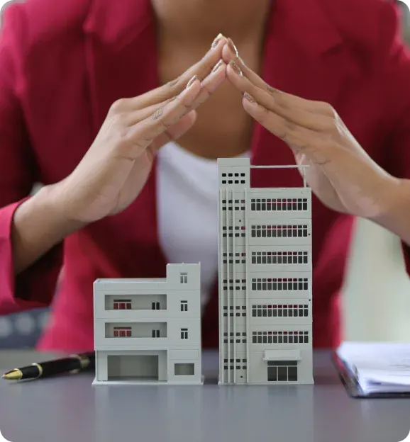 Person in a red jacket forming a protective gesture over two small architectural building models on a table.