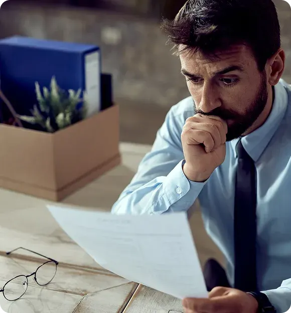 Worried man in a dress shirt and tie reading a document at a desk with a box of personal items and glasses nearby.