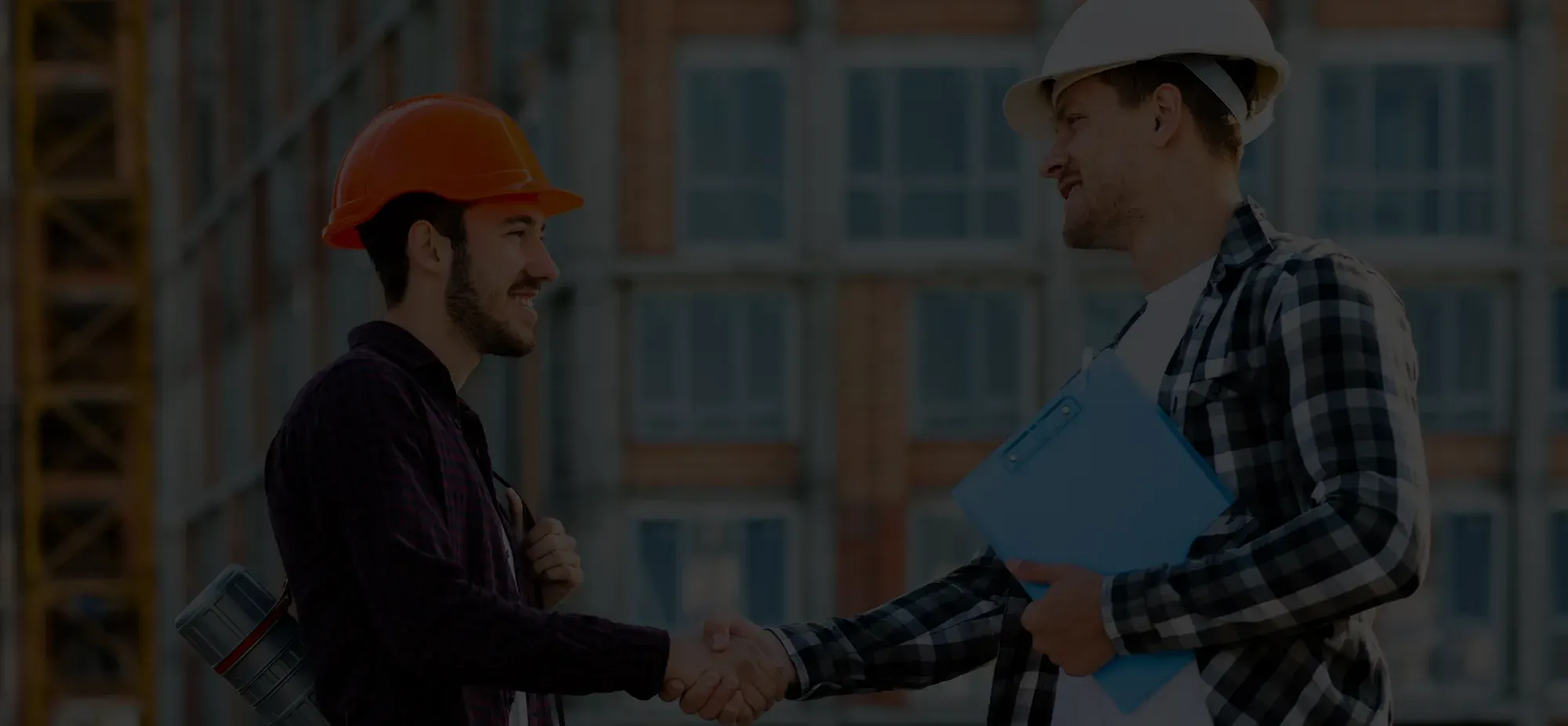 Two construction workers wearing hard hats shaking hands at a building site, one holding a blue clipboard.