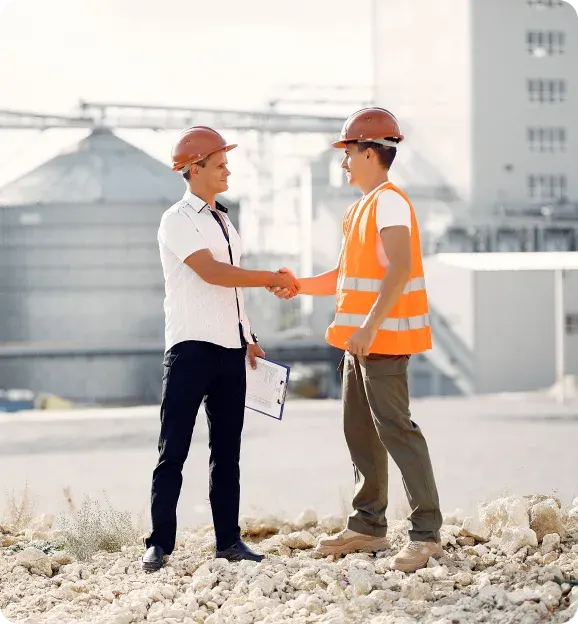 Two construction workers wearing helmets shaking hands outdoors near an industrial site with one holding a clipboard.