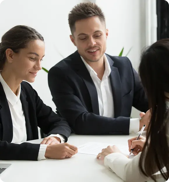 Two business professionals sitting at a table while a third person signs a document.