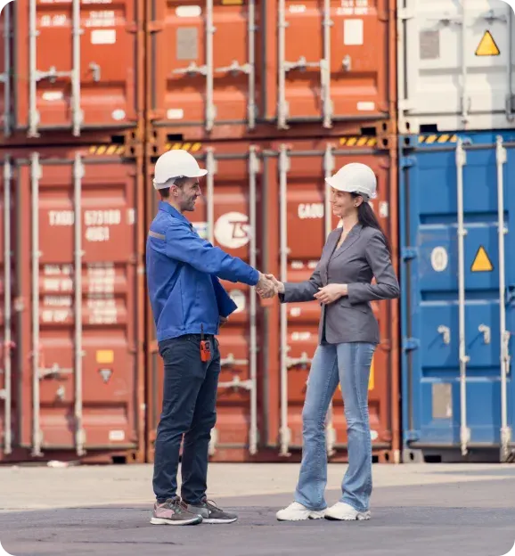 Two logistics workers wearing white hard hats shaking hands in front of stacked red and blue shipping containers.