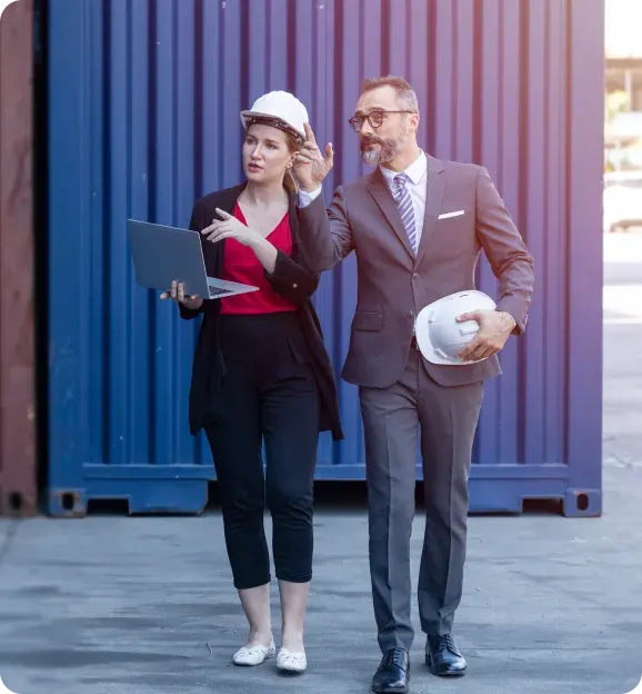 Two professionals, a woman with a white hard hat holding a laptop and a man in a suit holding a hard hat, discussing in front of a blue shipping container.