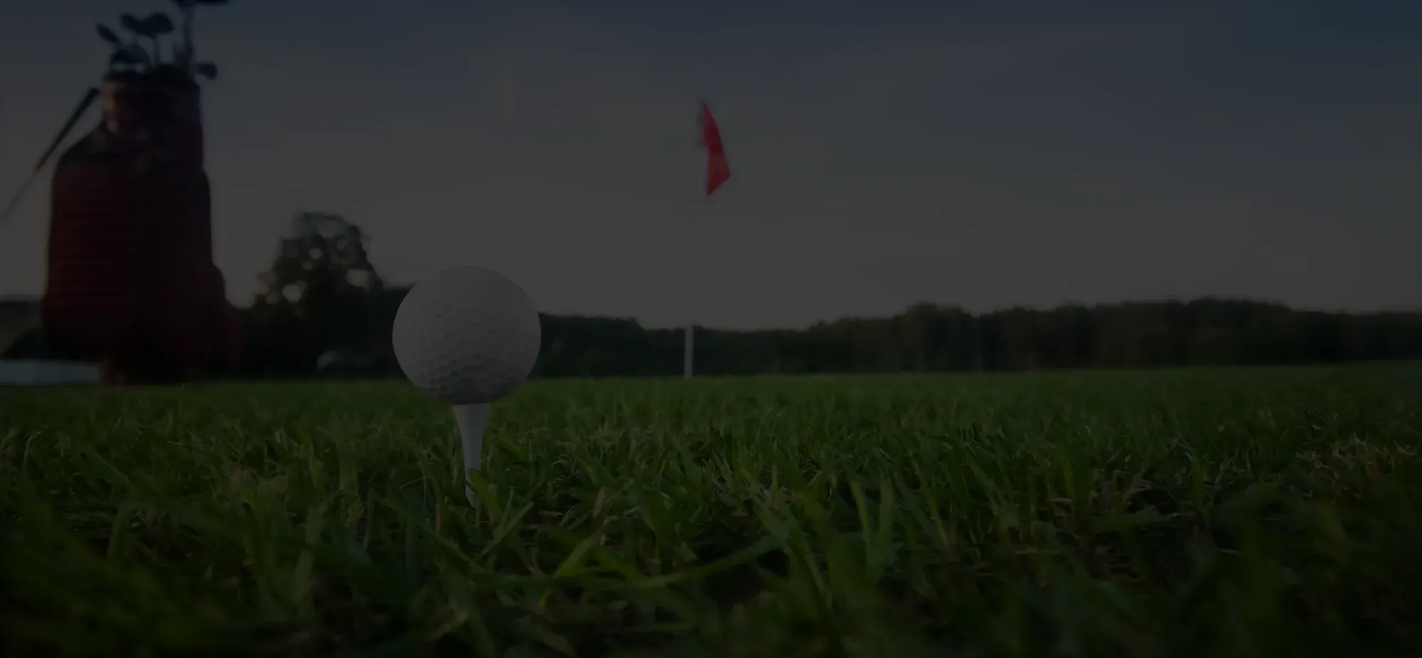 Close-up of a golf ball on a tee on green grass with a red flag and golf bag in the background.
