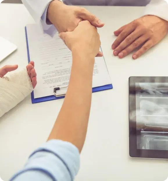 Patient with a bandaged hand shaking hands with a doctor over a clipboard with medical documents and an X-ray image on a tablet.