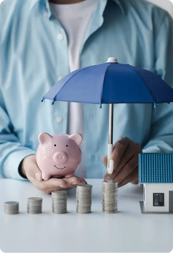 Person holding a pink piggy bank and a blue umbrella shielding stacks of coins and a small house model on a table.