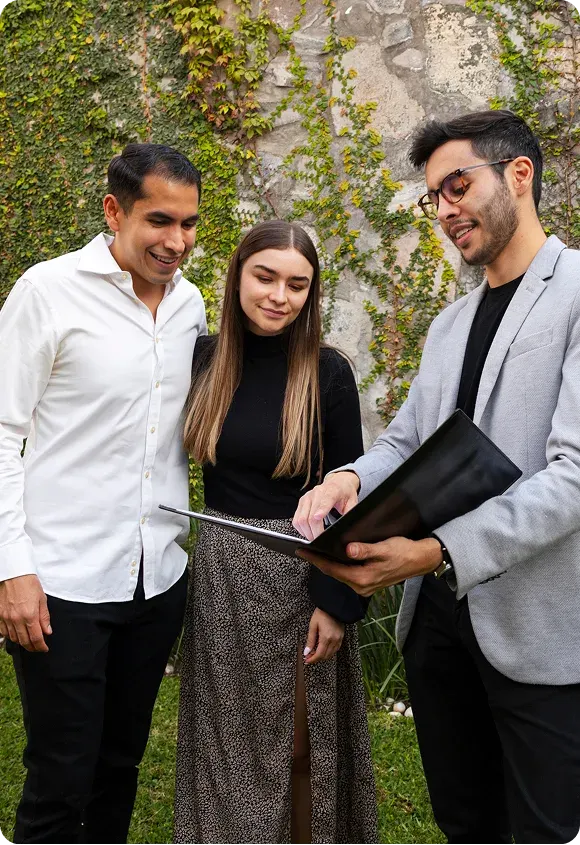Three young professionals standing outside in front of a ivy-covered wall, looking at documents in a black folder.