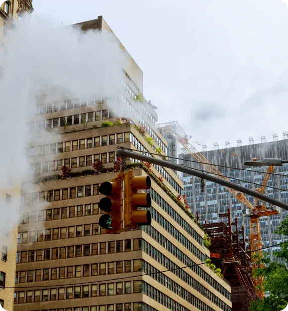 Urban scene with a traffic light showing green, steam rising near a tall building with terraces of plants, and cranes in the background.