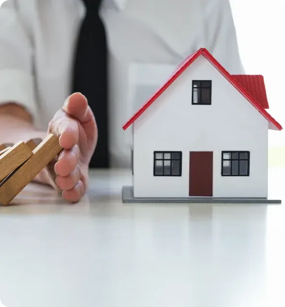 Person in a shirt and tie stopping falling wooden blocks next to a small model house with a red roof.