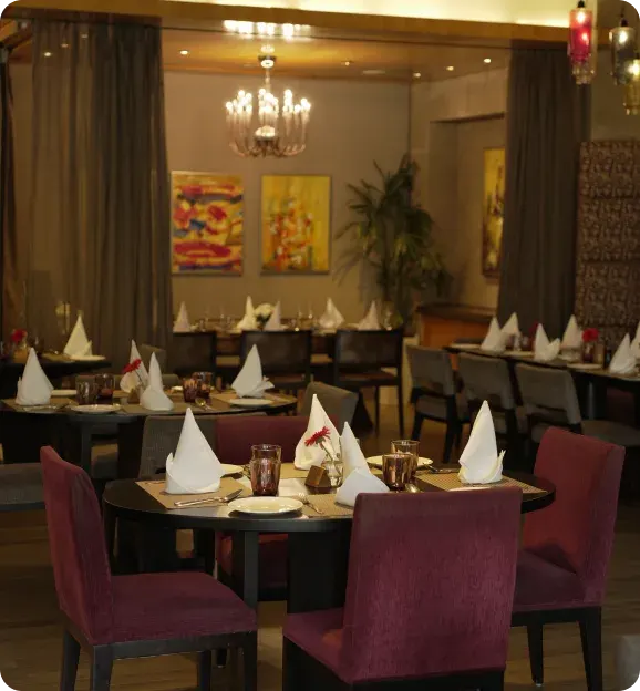 Cozy restaurant dining area with round tables set with white napkins, brown glasses, and pink chairs under warm lighting and abstract wall paintings.
