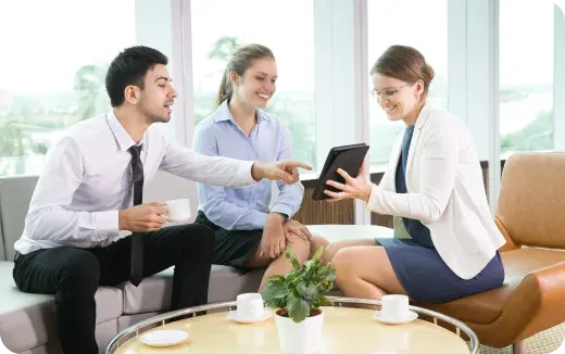 Three business professionals sitting and smiling, one pointing at a tablet held by a woman in a white blazer.