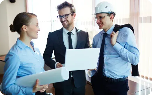 Three professionals discussing a project, including a woman holding blueprints, a man in a suit with a laptop, and a man wearing a hard hat holding a jacket over his shoulder.