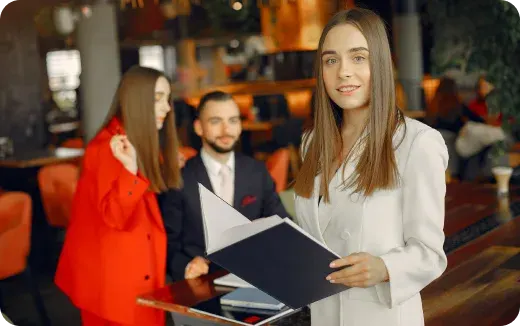 Three young professionals in a modern office; a woman in white holding a folder in the foreground, with a seated man and a woman in red in the background.