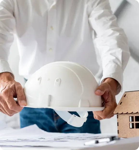 Person in white shirt holding a white construction hard hat over architectural blueprints with a small wooden house model on the table.