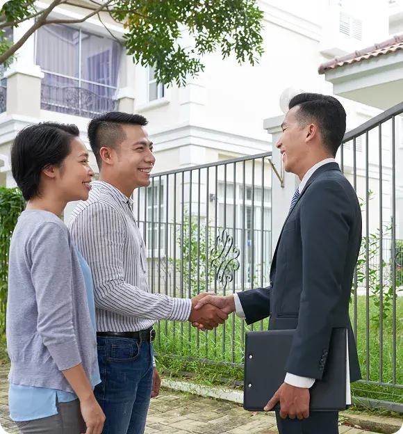 Smiling couple shaking hands with a man in a suit holding a folder outside a gated house.