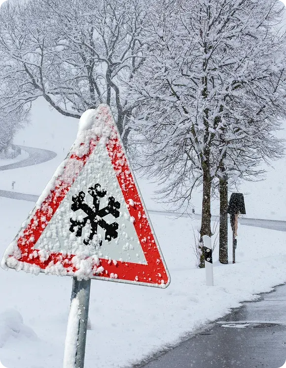 Snow-covered road sign with a black snowflake symbol warning of icy conditions, with a snowy, winding road and leafless trees in the background.