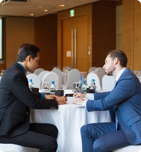 Two men in business suits seated at a table with bottled water and glasses, engaged in a discussion in a conference room.