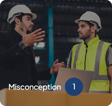 Two male workers wearing white safety helmets discussing in a warehouse, one in a black suit gesturing hands, the other in a yellow safety vest listening.