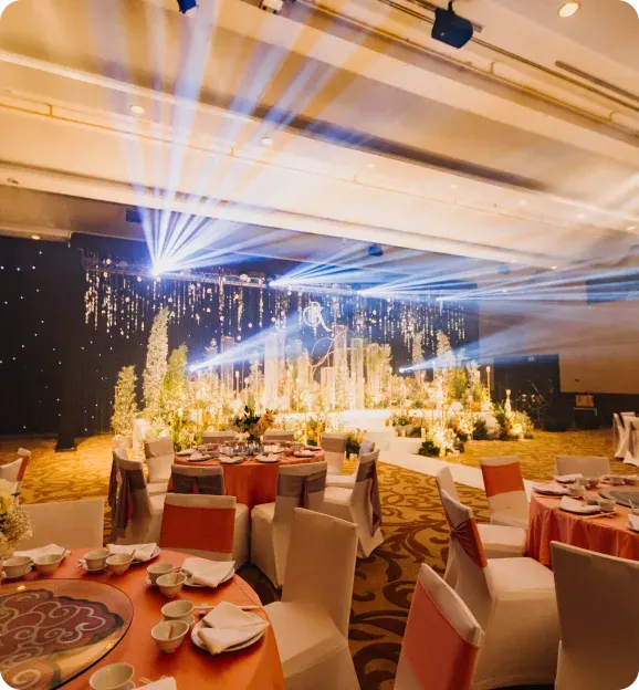 Indoor banquet hall with round tables covered in orange tablecloths and white chairs with orange sashes, decorated stage with plants and lights, and bright spotlights shining from the ceiling.