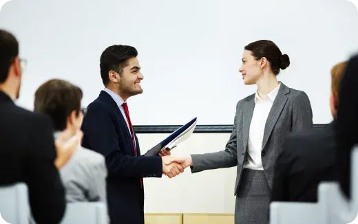 Professional man and woman shaking hands in a business setting with an audience watching.