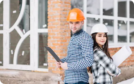 Man and woman wearing hard hats and checkered shirts standing back-to-back outside a building with rolled-up blueprints and clipboard.