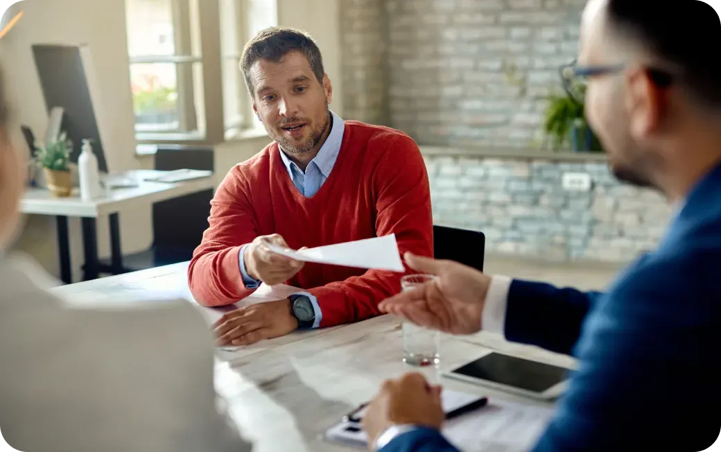 Man in red sweater handing a document to another person during a meeting in a modern office.