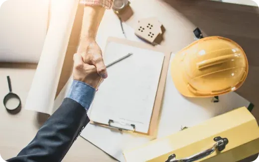 Two people shaking hands over a desk with a clipboard, yellow hard hat, measuring tape, and architectural tools.