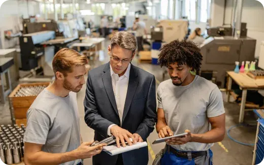 A man in a suit discusses a document with two workers holding tablets inside a manufacturing facility.