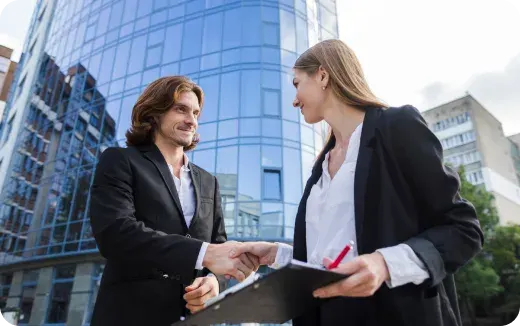 Man and woman in business attire shaking hands outside a modern glass office building.