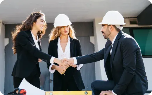 Two women and a man wearing business attire and white safety helmets shaking hands at a construction site.