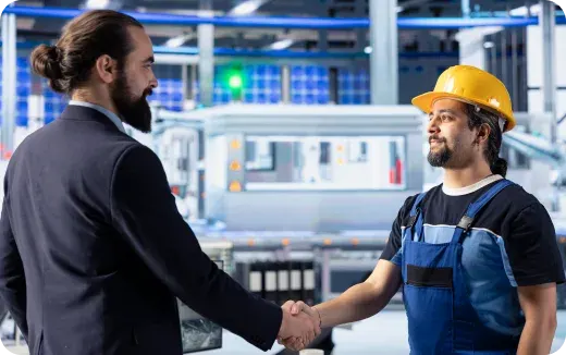 A businessman in a suit shaking hands with a factory worker wearing a yellow hard hat and blue overalls in an industrial setting.
