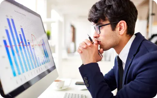 Man in a suit and glasses thoughtfully analyzing data charts on a computer screen in an office.