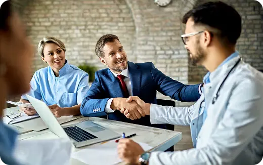 Businessman shaking hands with a doctor across a table while another medical professional watches and smiles.
