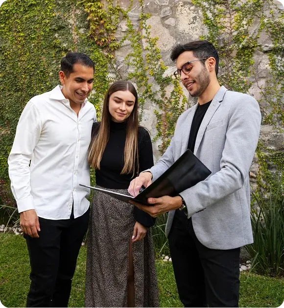 Three people standing outdoors in front of a vine-covered stone wall, one man in a light gray blazer showing a folder to a man in a white shirt and a woman in a black top and patterned skirt.
