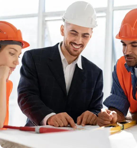 Three construction professionals, two wearing orange helmets and one in a white helmet and suit, reviewing building plans together.