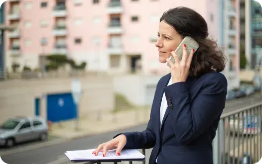 Businesswoman in a navy blazer talking on a smartphone while holding documents outdoors near a railing.