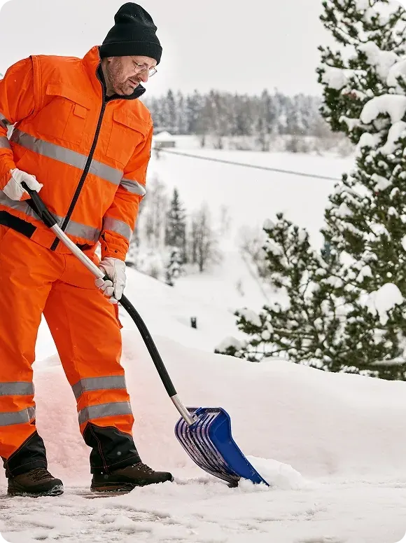 Man in bright orange winter workwear shoveling snow with a blue snow shovel on a snowy surface, with snow-covered trees and a winter landscape in the background.