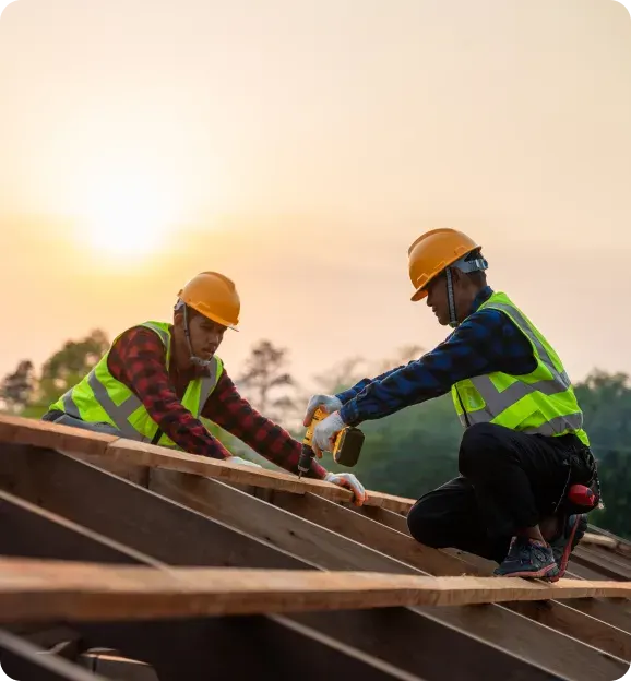 Two construction workers in safety vests and helmets working on wooden roof framing during sunset.