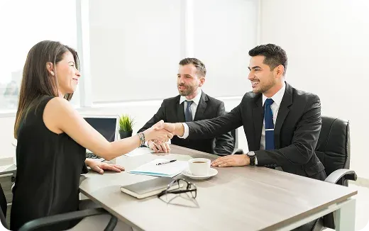 Two businessmen in suits shaking hands with a woman across a table in a bright office.