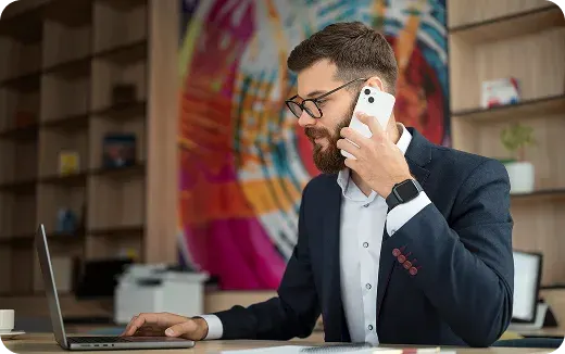 Bearded man wearing glasses and a suit jacket talking on a smartphone while using a laptop in a modern office.
