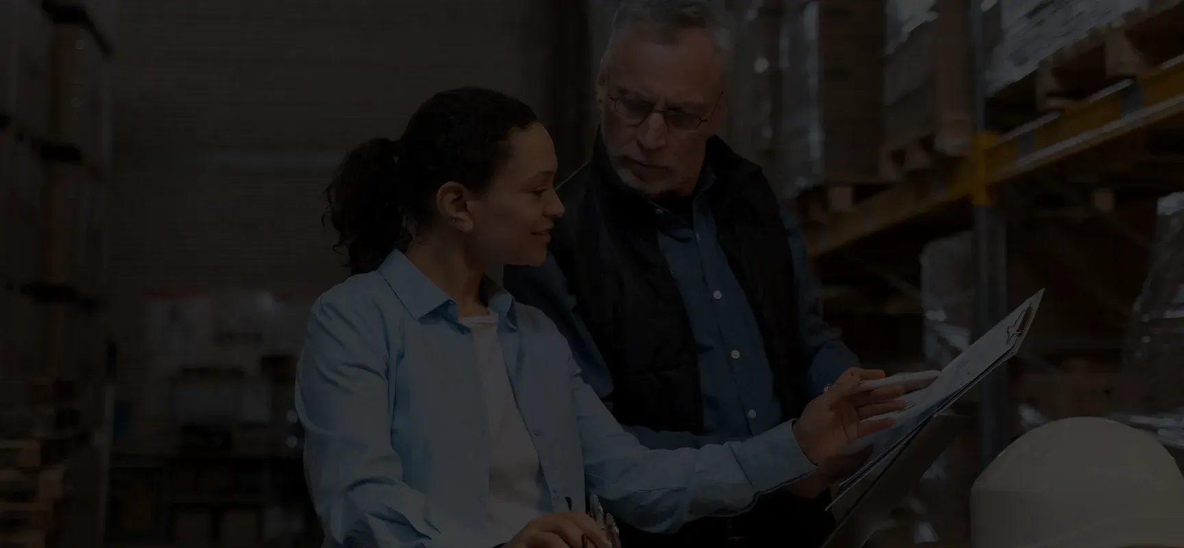 Two warehouse workers reviewing documents on a clipboard among stacked pallets and shelves.