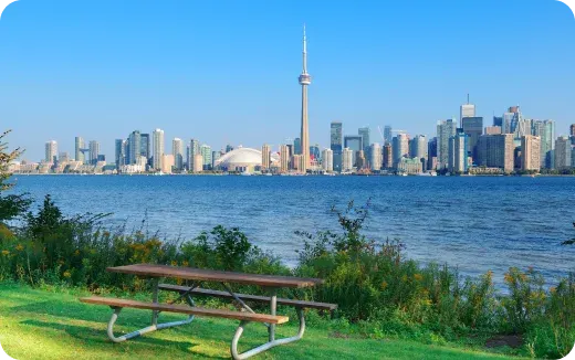 Picnic table on grassy shore with green bushes overlooking Toronto skyline and CN Tower across the water under clear blue sky.