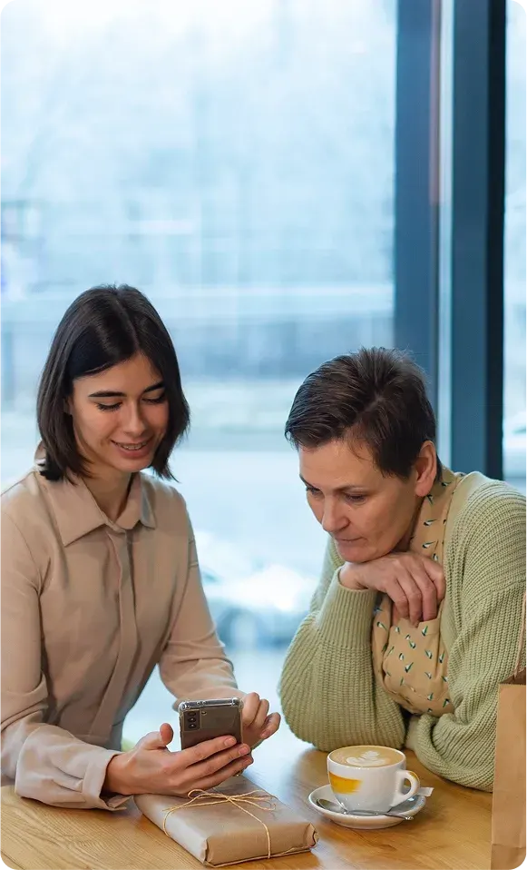 Two women sitting at a wooden table, one showing something on her smartphone to the other, with a cup of coffee and a wrapped gift on the table.