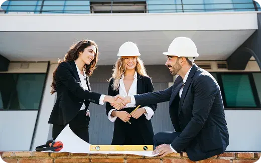 Two businesswomen and a businessman wearing white hard hats shaking hands at a construction site with building plans and measuring tools on a brick wall.