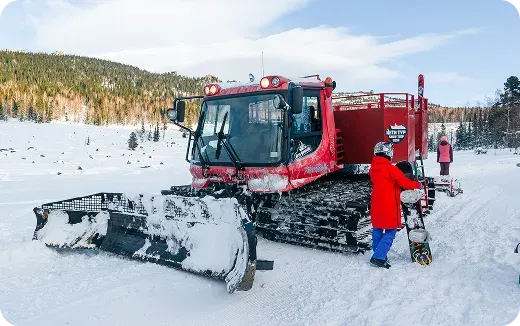 Red snow grooming machine parked on snow with two people in winter gear, one holding a snowboard.